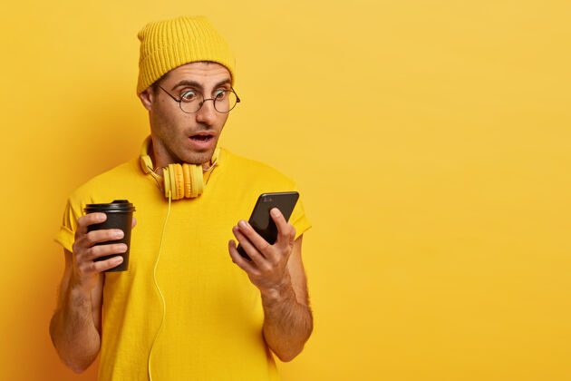 头饰惊讶的男人盯着智能手机 戴着透明眼镜和黄色帽子 喝着外卖咖啡 惊讶于互联网上的可怕消息单色 黄色耳机惊喜姿势