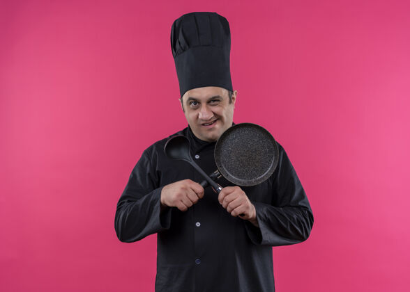 站着男厨师身穿黑色制服 头戴厨师帽 手拿煎锅和勺子 双手交叉 站在粉色背景上微笑着看着镜头拿着穿交叉