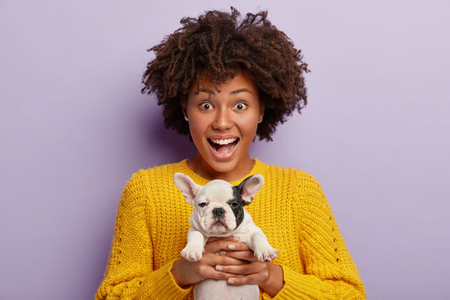 友谊快乐的非裔美国宠物主人照片抱着一只黑白相间的小狗 有着快乐的表情 穿着黄色的针织毛衣 关心宠物 想买什么产品来获得健康的营养黄色站微笑
