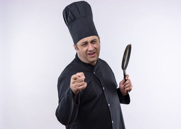 帽子男厨师身穿黑色制服 头戴厨师帽 拿着平底锅 手指指着镜头 快乐而积极地站在白色背景上厨师指着白