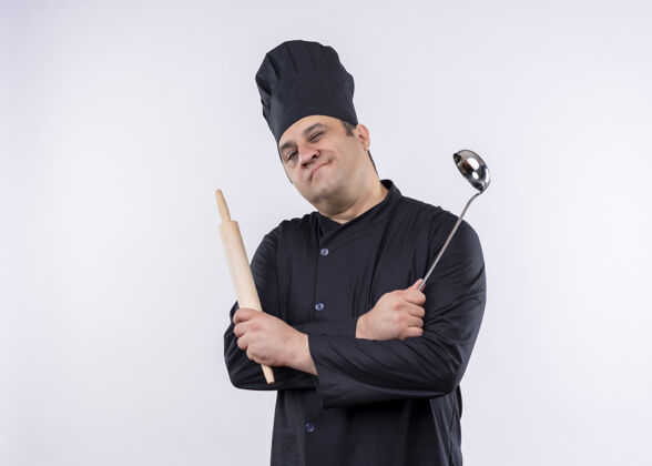 制服男厨师身穿黑色制服 头戴厨师帽 拿着勺子和擀面杖 微笑着看着站在白色背景上的摄像机黑帽子眨眼
