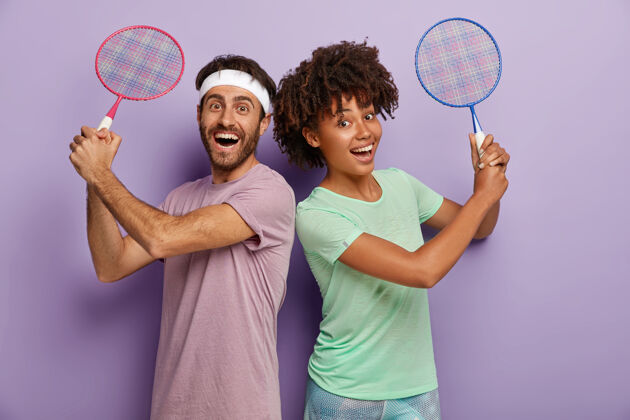 情侣快乐的混血男女背对背站着 握着网球拍 享受打球的乐趣 开怀大笑 身着t恤 积极而满意休闲美国人快乐