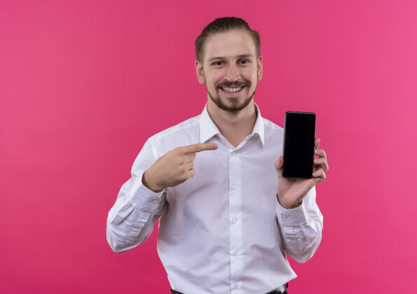 指点身着白衬衫的帅哥商人展示智能手机 手指指着智能手机微笑 粉色背景下站着一张幸福的脸秀白色帅气