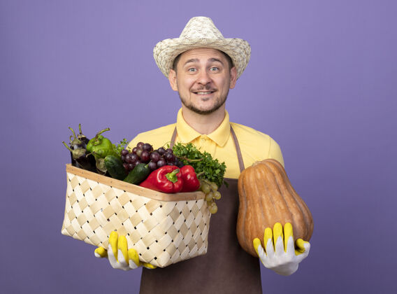 南瓜年轻的园丁 穿着连体衣 戴着帽子 戴着工作手套 手里拿着装满蔬菜的箱子 南瓜满脸笑容满的男人工作