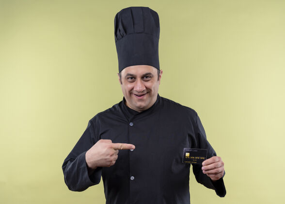 男性男厨师身穿黑色制服 头戴厨师帽 手拿信用卡 手指指着信用卡 站在绿色背景上开心地笑着绿色欢呼厨师