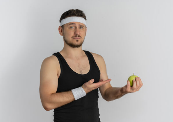苹果戴着头巾拿着青苹果的年轻健身男子用胳膊哦手看着严肃的脸站在白色的背景上抱着脸站着