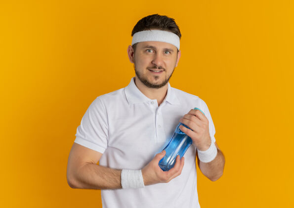 白色身穿白衬衫 头箍着水瓶 脸上带着微笑的年轻健身男子站在橙色背景下看着摄像机微笑橙色头带