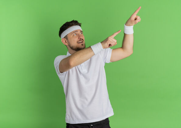 白色身穿白衬衫的年轻健身男子戴着头巾 一边看一边用食指指着站在绿色背景上的一边壁板手指看