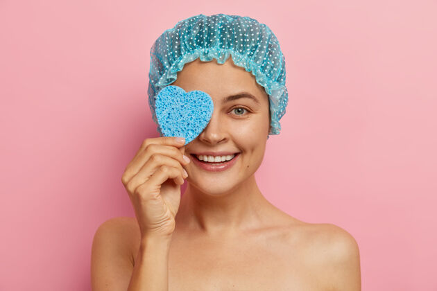 裸体积极快乐的可爱女人用心形海绵遮住眼睛 擦干净脸 戴上蓝色的保护性淋浴帽 接受美容治疗 对着粉色的墙壁做模特呵护和放松愉悦女人年轻