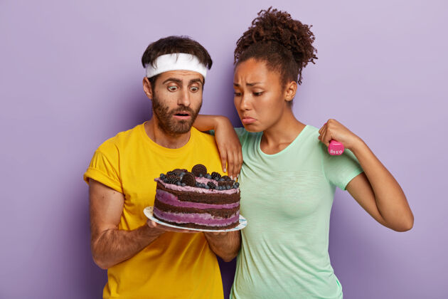 惊喜不快乐的运动型多样化的年轻男女带着诱惑看着美味的蛋糕 想吃却意识到它的危害 带着哑铃训练 穿着休闲装不满意美味女士