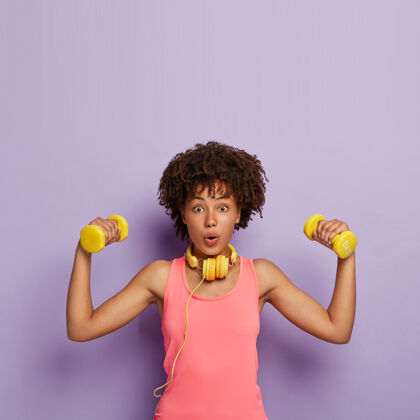 运动减肥和锻炼的概念惊讶的黑皮肤女人卷发 举哑铃 训练肌肉 有简单的肱二头肌锻炼 穿着休闲的粉红色上衣 使用耳机力量紫色Omg