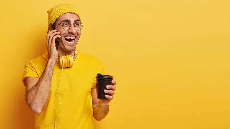 男性快乐的年轻人通过手机聊天 拿着外卖咖啡 喜欢喝酒 穿着休闲t恤和帽子设备电子咖啡
