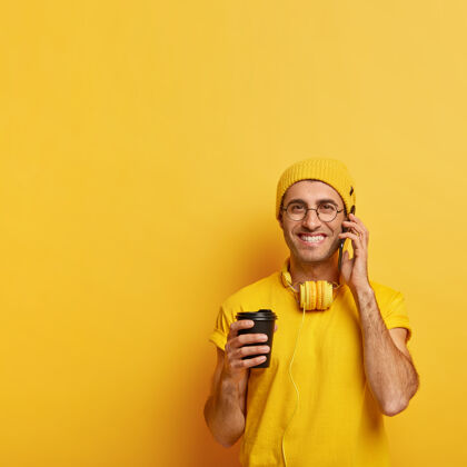 数码高兴的男模特通过手机给朋友打电话 拿着手机 一边喝外卖咖啡一边聊天 穿着黄色衣服 戴着透明眼镜欢呼黄色眼镜