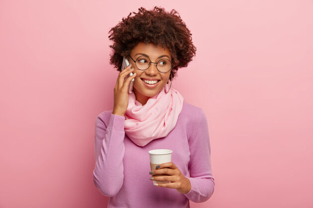 罗西积极的黑皮肤女人 留着非洲式发型 通过手机交谈 拿着外卖咖啡 享受愉快的交谈 穿着休闲套头衫和丝巾 笑容灿烂 讨论一些事情女人紫罗兰欢呼