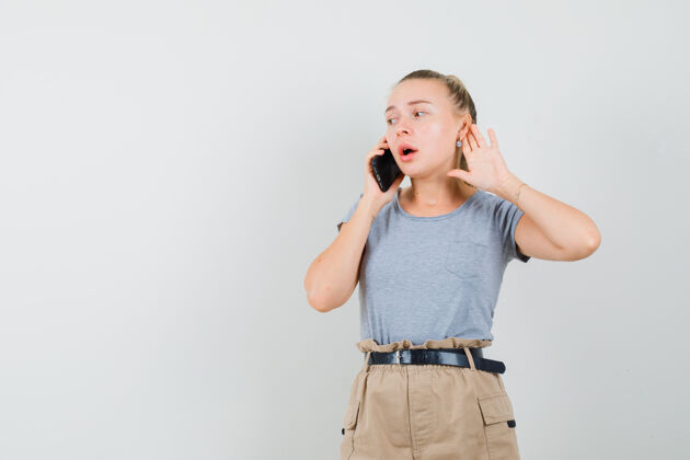 持有年轻的女性在讲手机 手放在耳后 穿着t恤 裤子 正面照人时尚电话