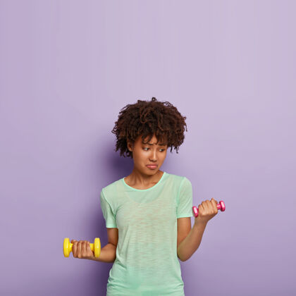 垫子照片中不满的黑皮肤女人举起哑铃 正在锻炼 想要强壮健康 穿着休闲t恤 举重活动运动装紫色