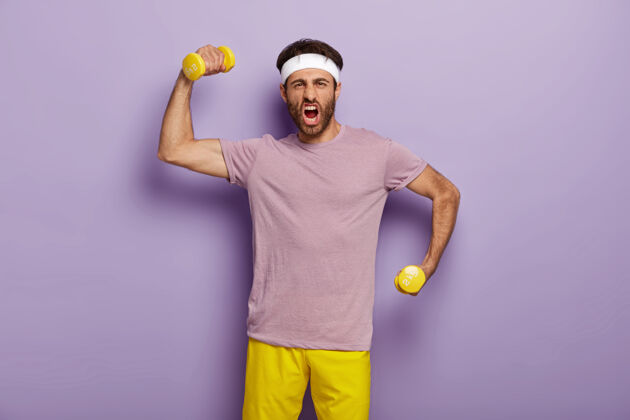健身房有趣的运动员举着哑铃 情绪化地喊叫 感觉强壮和运动 穿着紫色t恤和黄色短裤 站在室内男人在健身房锻炼 做运动健美紫色微笑哑铃