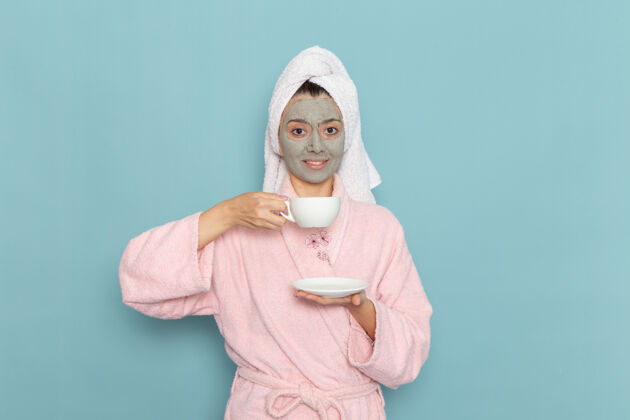 饮用正面图身着粉色浴袍的年轻女性淋浴后 微笑着喝咖啡 在蓝色墙壁上清洁美丽干净的水 自我护理的奶油淋浴浴袍年轻人自理