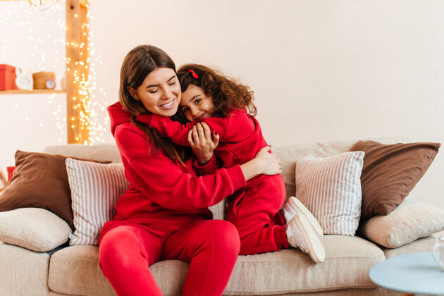 健康年轻的母亲坐在沙发上和一个未成年的女孩在一起室内拍摄的是穿着红色衣服的妈妈和女儿在沙发上拥抱的镜头房子女儿沙发