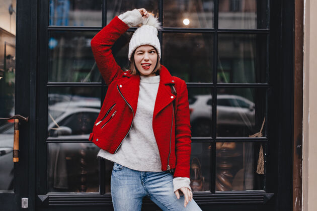 肖像穿着红色外套和牛仔裤的可爱女孩玩得很开心 眨着眼睛 露出舌头 把针织帽子蓬蓬靠在窗户上地图城市温暖