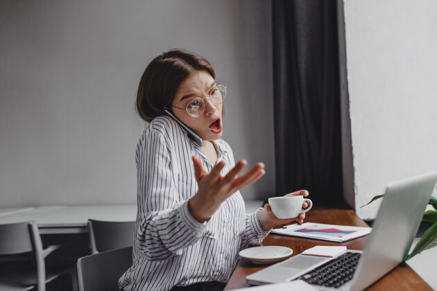 笔记本电脑戴眼镜的女工情绪激动 愤愤不平地打电话 坐在办公室里拿着笔记本电脑和一杯咖啡年轻人互联网电话