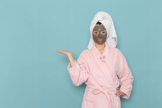年轻人正面图身着粉色浴袍 脸上戴着面具的年轻女性在蓝色墙壁上沐浴清洁美容自护霜伪装面霜脸