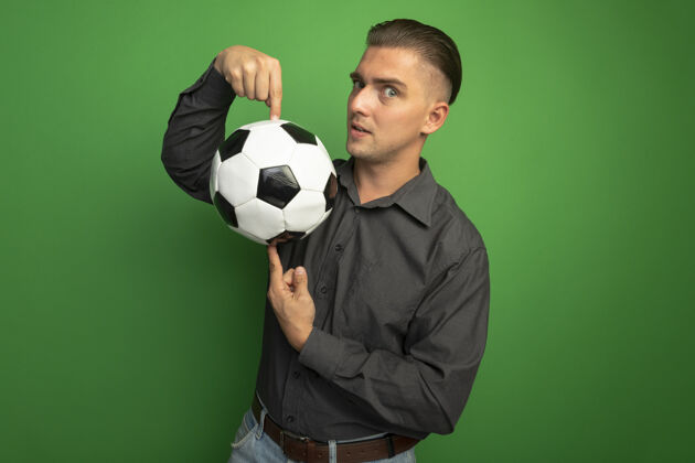 年轻身穿灰色衬衫的年轻帅哥展示着足球 食指指着球微笑着自信地站在绿色的墙上灰色指向自信