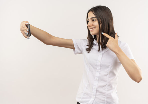 手机穿着白衬衫的年轻女孩用手机自拍 微笑着自信地站在白墙上拿着公民自信