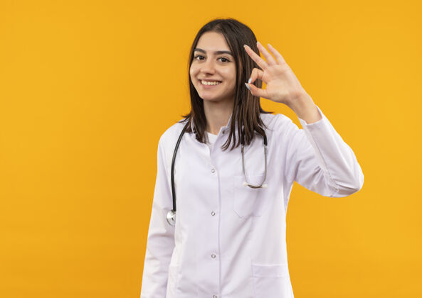 公民身穿白大褂的年轻女医生脖子上戴着听诊器 手指微笑着站在橙色的墙上做着“ok”的手势医院脖子办公室