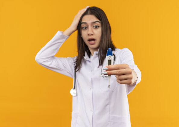 手势年轻的女医生穿着白大褂 脖子上戴着听诊器 显示着数字体温计 站在橙色的墙上惊讶地看着它周围站姿势