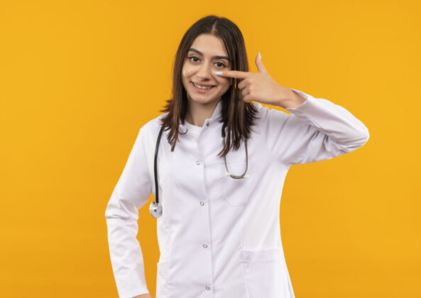 女性年轻的女医生穿着白大褂 脖子上戴着听诊器 手指指着眼睛 站在橙色的墙上微笑着脖子姿势人