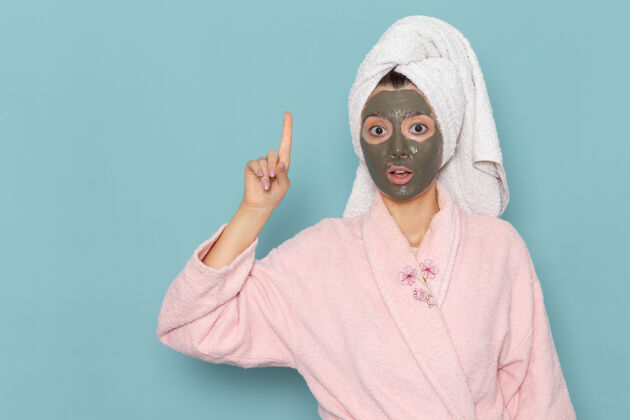 自我护理正面图身穿粉色浴袍 脸上戴着面具的年轻女性在蓝色墙壁上摆姿势沐浴清洁美容自护霜视野正面淋浴