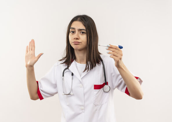 医生年轻的女医生穿着白大褂 脖子上戴着听诊器 手拿着数字体温计 站在白墙上严肃的脸上做着停止的手势手势做听诊器