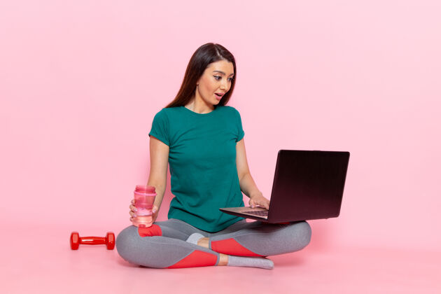 便携式电脑前视图身着绿色t恤的年轻女性在粉色墙上使用笔记本电脑腰部运动锻炼美丽苗条女性运动腰部工作电脑