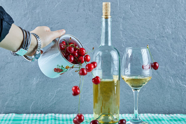 自然女人手上扔着一杯樱桃和一瓶白葡萄酒 杯子放在蓝色的表面上浆果饮料成熟