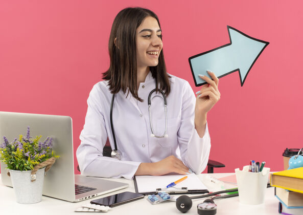 脖子年轻的女医生穿着白大褂 脖子上戴着听诊器 手里拿着蓝色的箭头 面带微笑地坐在桌边 粉色的墙上挂着笔记本电脑医院人外套