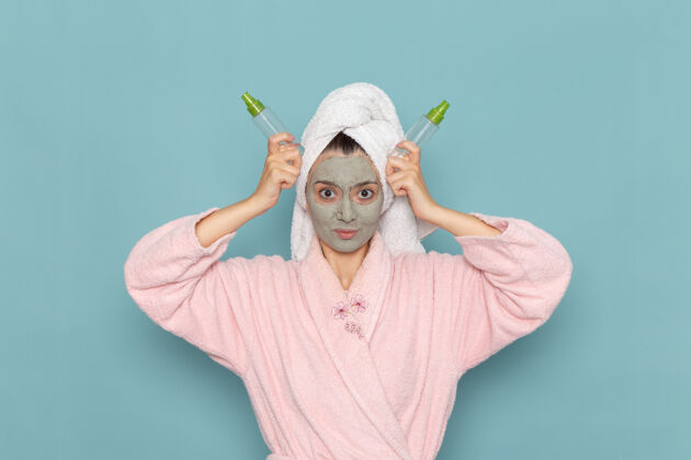 清洁正面图身穿粉色浴袍的年轻女性 脸上戴着口罩 拿着蓝色墙壁上的喷雾剂淋浴 清洁美容自我护理霜覆盖自我护理保持