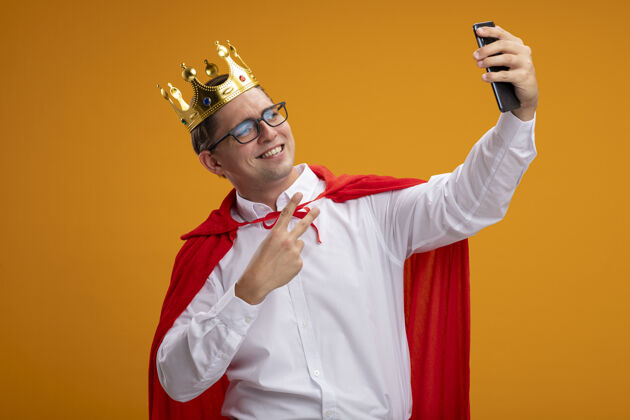 斗篷身穿红色斗篷 戴着皇冠眼镜的超级英雄商人用智能手机自拍 微笑着在橙色的墙上展示v形标志站立Vsign商务人士