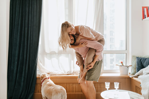 男朋友一个穿着轻装的男人背着他心爱的女朋友这对夫妇在他们的公寓里玩得很开心 而他们的狗正看着窗外欢笑拉布拉多情人