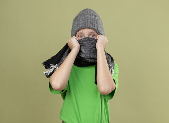 帽子生病的小男孩穿着绿色t恤 戴着暖和的围巾和帽子 戴着围巾 站在明亮的墙上 他看起来很担心围巾男孩温暖