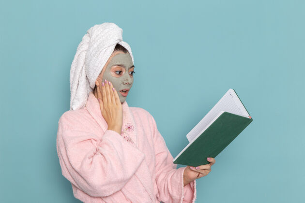 肖像前视图穿着粉色浴袍的年轻女性淋浴后阅读蓝色墙壁上的绿色抄写本美容水霜自助淋浴浴室抄写本阅读后面