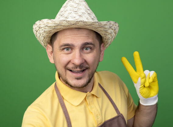 年轻年轻的园丁穿着连体衣 戴着帽子 戴着工作手套 面带微笑地看着前面 绿色的墙上挂着v字标志微笑快乐帽子