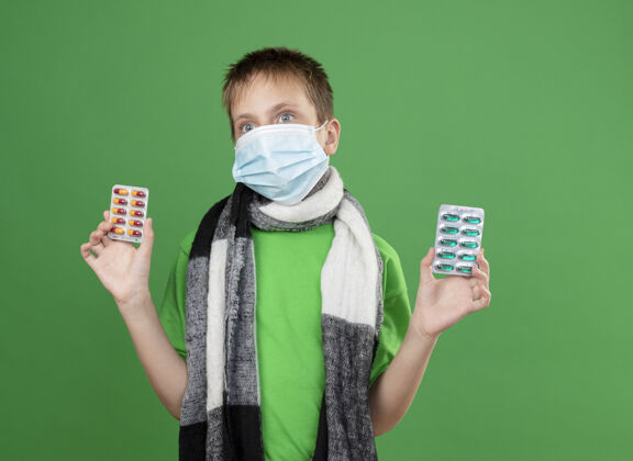 看着生病的小男孩穿着绿色t恤 围着暖和的围巾 戴着护面面具 手里拿着药片 站在绿色的墙上男孩站着温暖