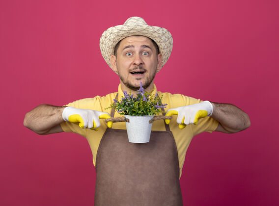 帽子年轻的园丁穿着连体衣 戴着帽子 戴着工作手套 手里拿着盆栽植物 站在粉红色的墙上 面带微笑地看着前方男人年轻脸
