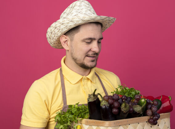 工人年轻的园丁穿着连体衣 戴着帽子 戴着工作手套 手里拿着装满蔬菜的箱子 站在粉色的墙上微笑着看着它们花园人人