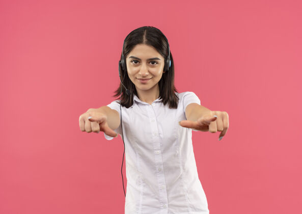 衬衫穿着白衬衫戴着耳机的年轻女孩 手指着前面微笑着自信地站在粉色的墙上手指公民麦克风