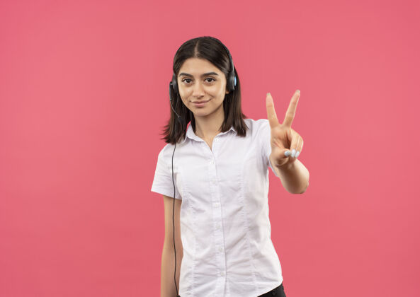 手势穿着白衬衫 戴着耳机的年轻女孩 手指着站在粉色墙上微笑的二号人物衬衫年轻人号码