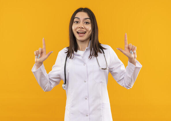 看年轻的女医生穿着白大褂 脖子上戴着听诊器 面带微笑地看着前面 食指站在橙色的墙上站外套目录