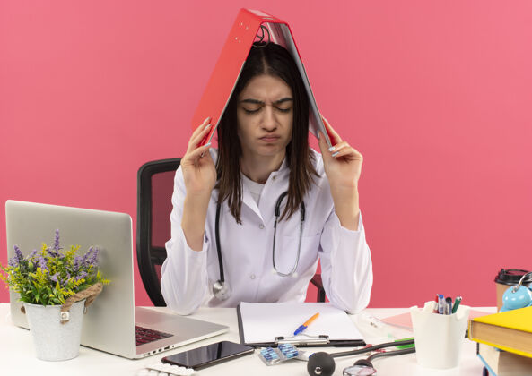 看年轻的女医生 穿着白大褂 脖子上戴着听诊器 头上戴着文件夹 坐在桌边 粉色的墙上挂着笔记本电脑 看上去很疲惫 工作过度医生脖子女人