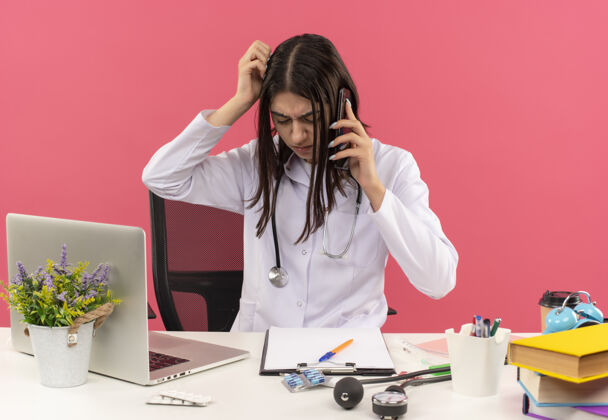 坐着年轻的女医生穿着白大褂 脖子上戴着听诊器 一边讲手机 一边神志不清地坐在桌旁用笔记本电脑在粉红色的墙上搔着头听诊器手机脖子